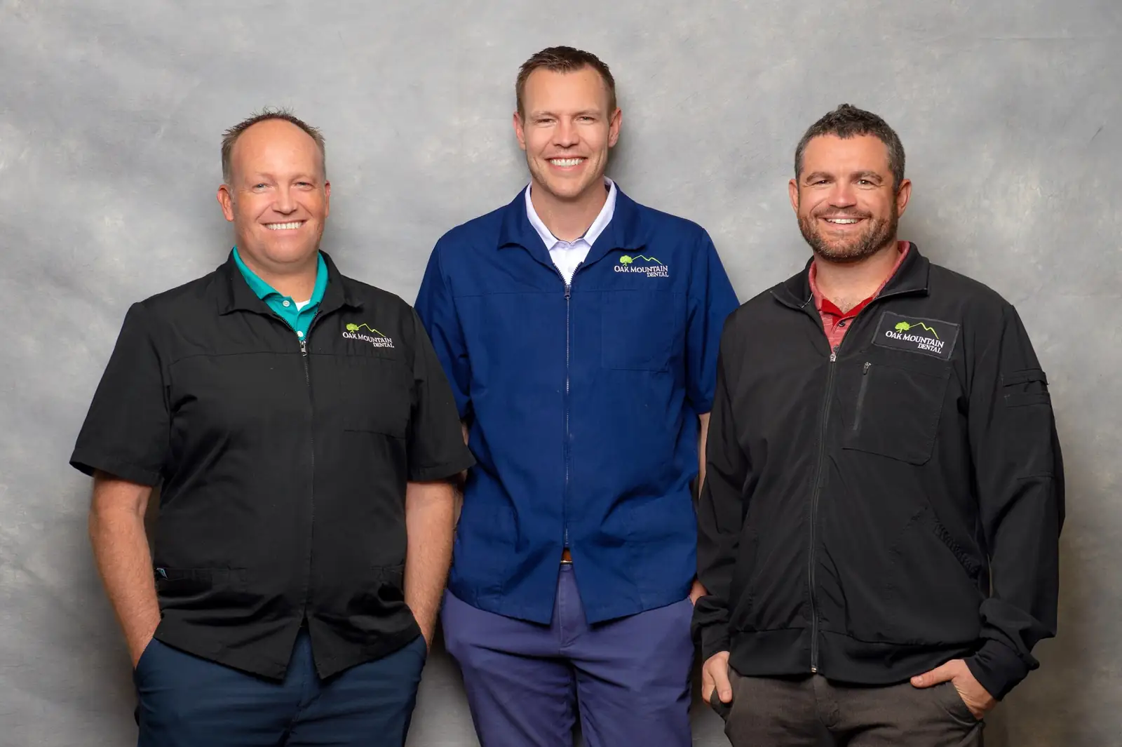 Three smiling dental professionals wearing Oak Mountain Dental uniforms in front of a gray backdrop.